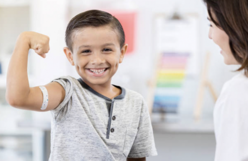 child holding up arm in flexed position with band aid on it with doctor looking on smiling